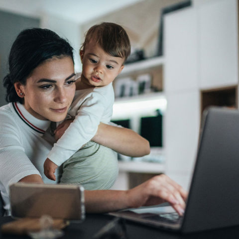 Woman holding toddler while working on laptop