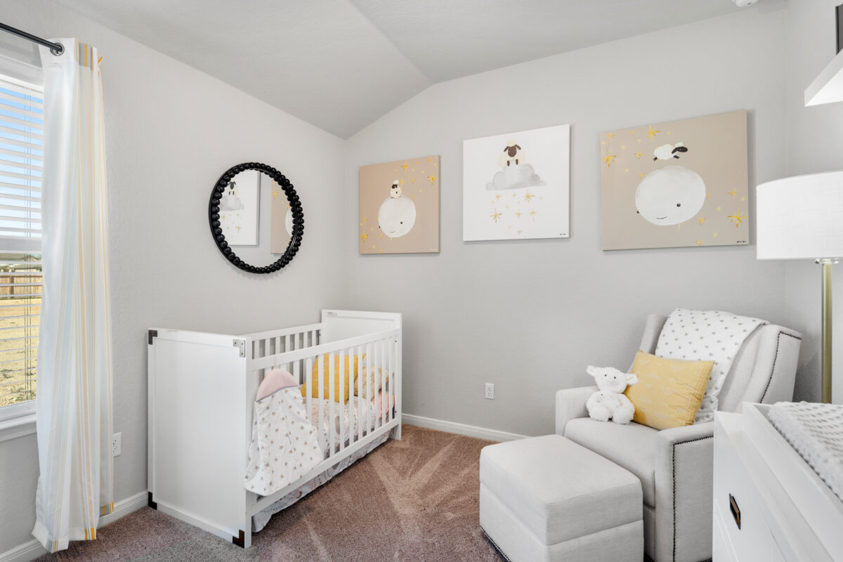 View of secondary bedroom furnished with crib, chair with ottoman and framed art.