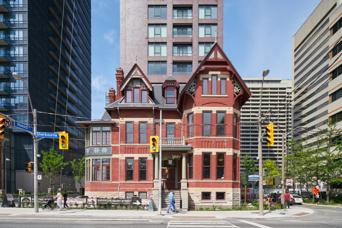 Exterior view of red brick Victorian building set among modern high-rise buildings in city center.