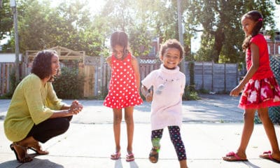 Smiling woman crouching looking at 3 young girls playing outdoors.