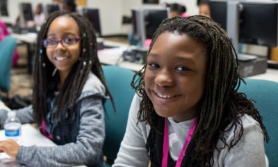 Two smiling young girls sitting at their desks in a computer lab.