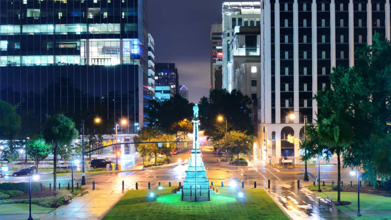 Downtown night scene. A monument in a park is the focus, and there are office buildings, streetlights and cars in the street.